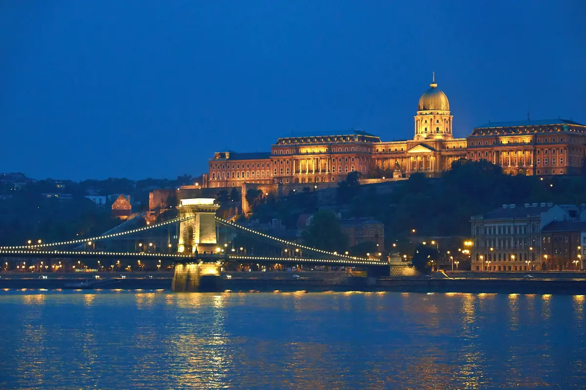 The Buda Castle palace and the Chain Bridge lit against a deep blue sky at dusk, seen across the Danube.