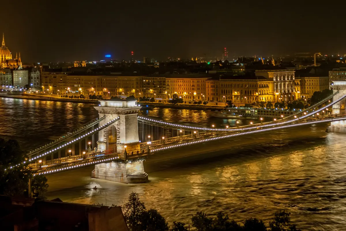 The Széchenyi Chain Bridge lit with strings of lights at night, its stone towers and the Pest embankment glowing in the background.