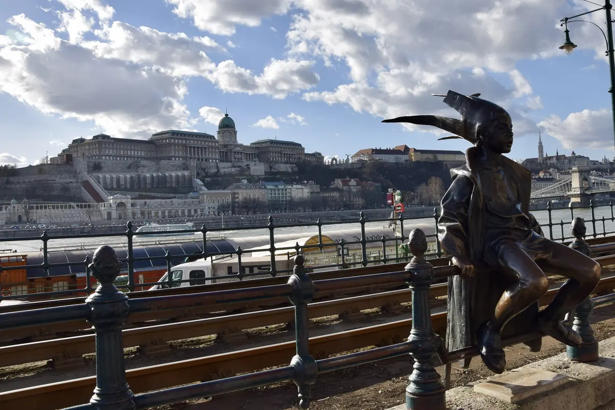 The Little Princess statue sitting on the railings of the Danube promenade, with the Buda Castle palace on the hill across the river.